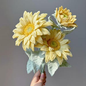 May include: A bouquet of artificial sunflowers with yellow petals and green centers. The leaves are a muted green color. The flowers are held in a hand against a gray background.