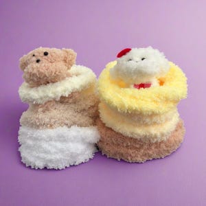 May include: Two plush toy figures, one a teddy bear and the other a cat, each atop a stack of fluffy, layered fabric. The teddy bear is light brown with a cream-colored collar and white base. The cat has a red bow and a red spot on its head.