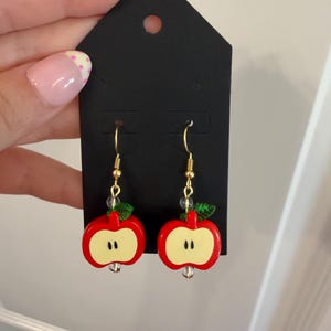May include: A pair of dangle earrings featuring red apple slice charms with a pale yellow center and two small black seeds. Each earring has a small green leaf accent, a clear bead, and a gold-toned metal hook for pierced ears.