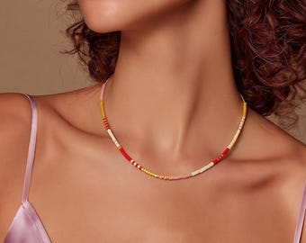 May include: A delicate beaded necklace featuring a repeating pattern of small, cylindrical beads in shades of soft pink, bright yellow, vibrant red, and cream. The necklace rests against the neck, showcasing a simple, lightweight design.