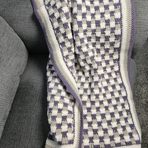 May include: A handmade crochet blanket in a checkerboard pattern of white, gray, and purple. The blanket has a border of white, gray, and purple. The blanket is draped over a gray couch.