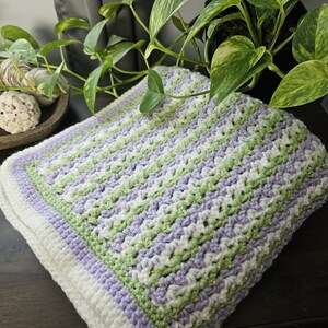 May include: A handmade crochet blanket in shades of white, light green, and lavender. The blanket features a textured stitch pattern with a white border. The blanket is folded and appears soft and cozy, suitable for a baby or as a decorative throw.