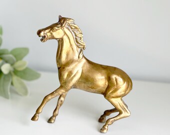 May include: A gold-colored metal horse figurine. The horse is standing on all four legs with its head turned to the side. The figurine is a decorative object.