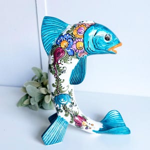 May include: A ceramic fish figurine featuring a white body with hand-painted floral motifs in shades of blue, orange, and pink. The fish has a bright turquoise head, fins, and tail, with a small opening at the top and an orange-rimmed mouth.