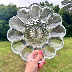 May include: A clear glass serving dish with a scalloped, flower-like edge and a central starburst pattern. The dish features a series of twelve rounded indentations around the perimeter, creating a petal-like appearance with a faceted texture.