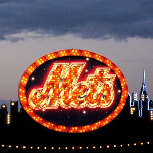 May include: A large, illuminated sign with the word "Mets" in a retro font, surrounded by a ring of lights. The sign is set against a silhouette of a city skyline under a cloudy sky. The sign is orange and red.