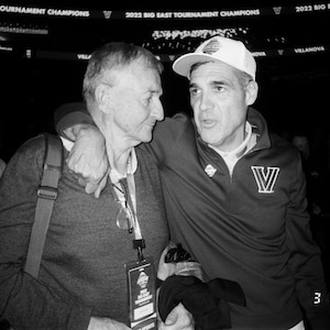 May include: Black and white photograph of two men embracing. One man wears a navy blue pullover with a white "V" logo and a white cap. The other man wears a gray sweater and glasses. The date "3 13 22" is visible.