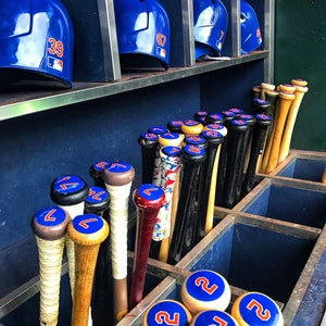May include: A close-up of a baseball equipment rack with blue and white baseball helmets and wooden baseball bats with blue and white numbers. The bats are stored in a rack with blue and white numbered caps.
