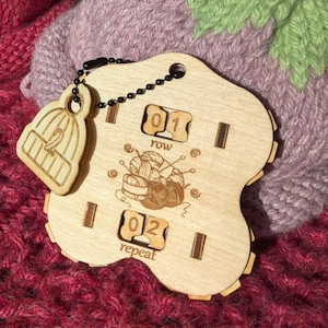 以下が含まれることがあります： 手作りのイチゴとナスを模した編み物のクローズアップ。イチゴは赤い色で緑の葉があり、ナスは紫で緑の葉があります。木製のステッチマーカーと行カウンターも表示されています。