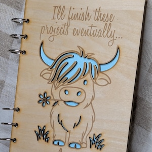 May include: Wooden notebook with a laser-cut design of a Highland cow with blue accents, holding a flower. The text "I'll finish these projects eventually..." is above the cow. The name "Anya" is below the cow. Other notebooks are visible.