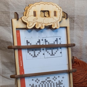 Puede incluir: Soporte de patrón de tejido de madera con diseño de oveja. El soporte muestra tres patrones de tejido con gráficos. Una madeja de hilo naranja y agujas de tejer están junto al soporte. El texto "Autumn Forest" y "Autumn Acorns" son visibles.
