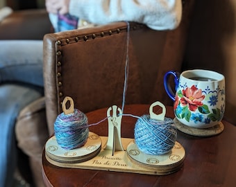 May include: A wooden yarn holder with two spools of colorful yarn, connected by a central guide. The base reads "Pirouette Pas de Deux". A floral mug of tea sits nearby, suggesting a cozy crafting scene.