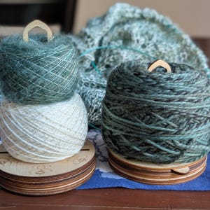 May include: Two skeins of yarn in shades of teal and cream, stacked on a wooden yarn holder. The yarn holders have a wooden handle. A partially visible knitted item is in the background.
