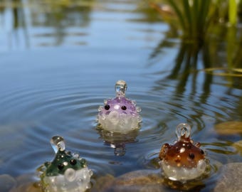 May include: Three colorful glass pufferfish figurines floating in water. One is green, one is purple, and one is brown. Each has a clear body with white bubble-like details and black eyes. The figurines are in a pond with rocks and greenery.