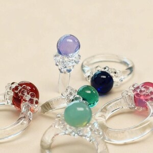 May include: A selection of clear glass rings, each with a colourful, spherical gemstone. Gemstones are in red, blue, green, and purple. The rings have a delicate, bubbly design around the gemstone.