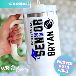 Könnte beinhalten: Weißer isolierter Becher mit Griff, mit dem Text "SENIOR 2026 BRYAN" in Schwarz, mit einer blauen Abschlusskappe und einer Football-Grafik. Der Becher hat auch den Text "Riverside High School" und "Wildcats".