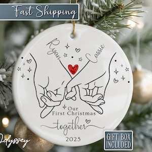 K&ouml;nnte beinhalten: Ein wei&szlig;er Keramik-Weihnachtsschmuck mit einer schwarzen Strichzeichnung von zwei H&auml;nden, die sich an den Fingern halten, einem roten Herzen und den Namen "Ryan" und "Cassie". Der Text "Our First Christmas together 2025" ist ebenfalls enthalten. Der Schmuck h&auml;ngt an einem Weihnachtsbaum.
