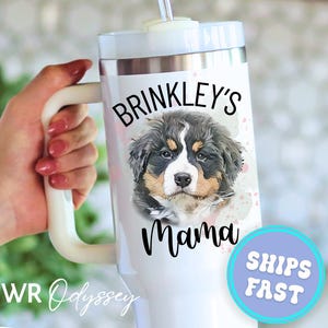 May include: White travel mug with a handle, featuring a portrait of a Bernese Mountain Dog puppy. The mug is labeled "Brinkley's Mama" in black script. A blue and purple circle with "SHIPS FAST" is also visible.