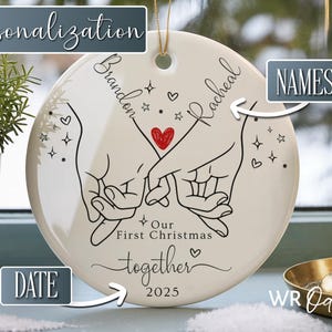K&ouml;nnte beinhalten: Wei&szlig;es Keramik-Weihnachtsornament mit goldener Aufh&auml;ngeschnur. Das Ornament zeigt eine schwarze Strichzeichnung von zwei H&auml;nden, die Finger halten, ein rotes Herz und die Worte "Our First Christmas together 2025".