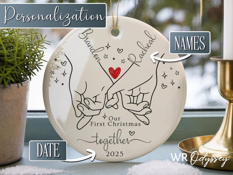 K&ouml;nnte beinhalten: Wei&szlig;es Keramik-Weihnachtsornament mit goldener Aufh&auml;ngeschnur. Das Ornament zeigt eine schwarze Strichzeichnung von zwei H&auml;nden, die Finger halten, ein rotes Herz und die Worte "Our First Christmas together 2025".