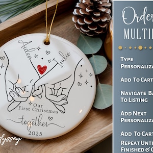 K&ouml;nnte beinhalten: Wei&szlig;er Keramik-Weihnachtsschmuck mit goldener Aufh&auml;ngeschnur. Das Ornament zeigt eine schwarze Strichzeichnung von zwei H&auml;nden, die sich an den Fingern halten, ein rotes Herz und die Namen "Aiden" und "Indie". Der Text lautet "Our First Christmas together 2025".