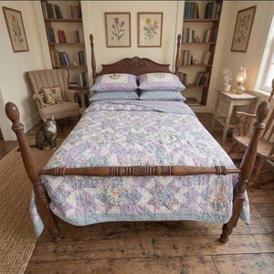May include: A wooden four-poster bed frame featuring a headboard with carved details, dressed in a quilt with a lavender, blue, and white star-patterned floral print, paired with two matching lavender pillowcases with floral embroidery.
