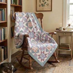 Peut inclure: Un quilt en patchwork avec un motif en étoile dans des tons lavande, bleu et blanc, agrémenté d'imprimés floraux et de petits animaux. Le quilt est drapé sur une chaise à bascule en bois, révélant un design géométrique avec une bordure bleu clair.