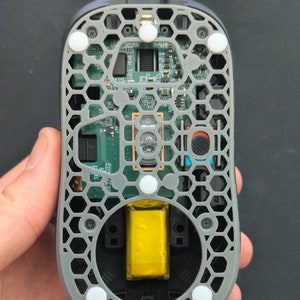 May include: The bottom of a gray computer mouse with a honeycomb pattern and a yellow battery visible inside. The mouse has five white rubber feet.