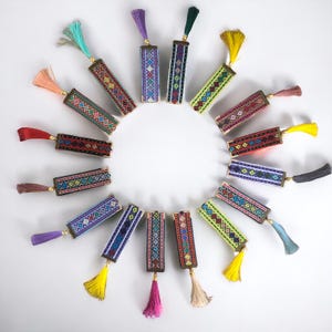 May include: A circle of 18 colourful tassel keychains with geometric patterns. Each keychain has a different colour tassel and a rectangular metal charm with a unique embroidered design.