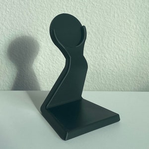 May include: A black plastic stand for holding a smart watch or other small device. The stand has a circular top and a curved base.