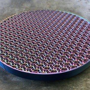 Puede incluir: Un posavasos redondo de metal con un acabado iridiscente de colores. El posavasos presenta un patrón repetitivo de bucles entrelazados, creando una superficie texturizada.