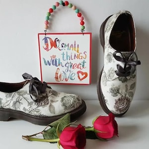 May include: Two pairs of white floral print shoes with black laces. The shoes have a brown sole and a white background with a floral pattern. The shoes are sitting on a white surface with a sign that says "Do small things with great love" in colorful letters.