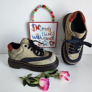 Puede incluir: Dos pares de zapatos de plataforma de cuero beige con ribetes negros y detalles rojos. Los zapatos tienen cordones y están atados. Los zapatos están sobre una superficie blanca con un letrero que dice "Do small things with great love" y una cuerda de cuentas de colores.