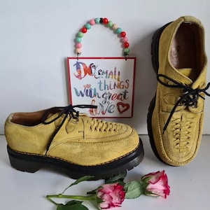 Può includere: Un paio di scarpe in pelle scamosciata gialla con lacci e suole spesse nere. Le scarpe hanno un motivo di cuciture decorative sulla punta e una chiusura con lacci neri. Le scarpe sono su una superficie bianca con un cartello che dice "Do small things with great love".