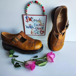 Può includere: Due paia di sandali in pelle marrone con chiusura a fibbia. I sandali hanno un cinturino sul piede e un cinturino alla caviglia. I sandali sono su una superficie bianca con un cartello che dice "Fai cose piccole con grande amore".