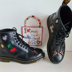 May include: A pair of black leather boots with laces and a variety of colourful patches, including cherries, a skull, and a butterfly. The boots have a thick, white sole. The text "Do small things with great love" is visible in the background.