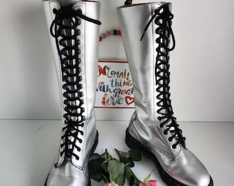 May include: A pair of silver knee-high boots with black laces. The boots have a shiny metallic finish and a classic lace-up design. The boots are displayed with pink roses and a sign that says "Small things with great love".