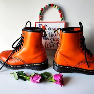 May include: A pair of orange leather boots with black laces and black soles. The boots are worn and have a distressed look. The boots are sitting on a white surface.