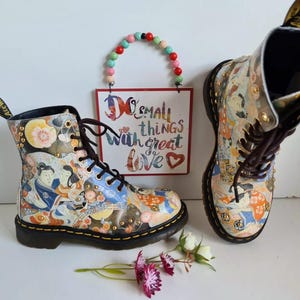 Könnte beinhalten: Ein Paar schwarze und weiße gemusterte Dr. Martens Stiefel mit goldenen Akzenten. Die Stiefel sind Schnürstiefel und haben eine dicke Sohle. Die Stiefel stehen auf einer weißen Oberfläche mit einem Schild, auf dem "Do small things with great love" steht.