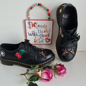 May include: A pair of black leather Dr. Martens shoes with embroidered patches. The patches include a cherry, a butterfly, and a spade. The shoes have yellow stitching and a black lace-up closure.