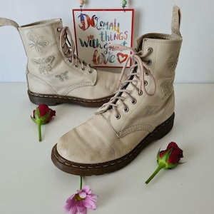 Puede incluir: Un par de botas de cuero de color blanco roto con cierre de cordones y bordados de mariposas. Las botas tienen una suela de goma marrón y están sobre una superficie blanca.