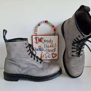 Può includere: Due stivali grigi con motivo a pelle di serpente, lacci e suole neri. Un piccolo cartello con la scritta "Do small things with great love" è sullo sfondo.