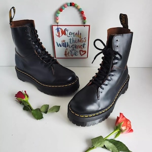 May include: A pair of black leather platform boots with yellow stitching and laces. The boots are on a white surface with two red roses.