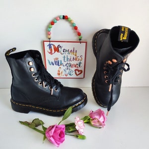 May include: Black leather ankle boots with gold eyelets and laces. The boots have a yellow stitching detail and a black and yellow Dr. Martens logo on the heel.