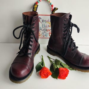 Puede incluir: Un par de botas de cuero burdeos con cordones negros. Las botas tienen un diseño de puntera y están sobre una suela negra. Dos rosas naranjas y un letrero con la frase "small things create love" están en el fondo.