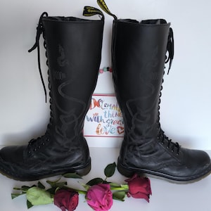 Peut inclure: Paire de bottes noires montantes à lacets, avec un motif serpentin sur le côté. Les bottes sont ornées d'une étiquette jaune et sont présentées avec des roses. Hauteur des bottes : environ 40 cm. Le signe dit "Small things with great love."