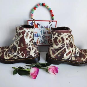 Könnte beinhalten: Ein Paar braune und weiße gemusterte Dr. Martens Stiefel mit schwarzen Schnürsenkeln. Die Stiefel haben einen weißen Hintergrund mit braunen und schwarzen abstrakten Mustern. Die Stiefel stehen auf einer weißen Oberfläche mit einem kleinen Schild, auf dem "No small thing with great love" steht.