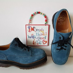 May include: A pair of blue suede shoes with black laces. The shoes have a thick, black rubber sole. The shoes are sitting on a white surface. The text on the sign reads "Do small things with great love."
