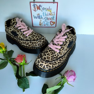 Può includere: Un paio di sneakers platform con stampa leopardata, suole nere e lacci rosa. Le scarpe sono esposte con rose e un cartello con la scritta "Do small things with great love."