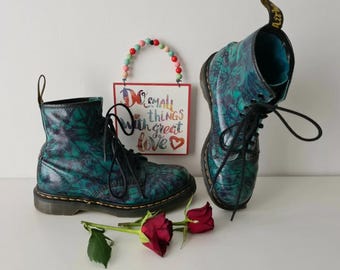 May include: A pair of teal blue and black Dr. Martens boots with a tie-dye pattern. The boots have black laces and are sitting on a white surface. The text "Do small things with great love" is visible on a sign behind the boots.
